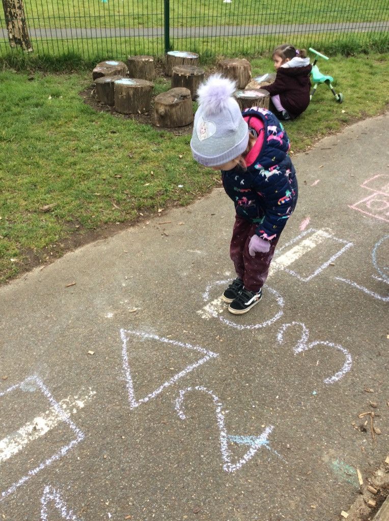 Child in a winter coat looking down at chalk drawings of numbers and shapes on a paved path.