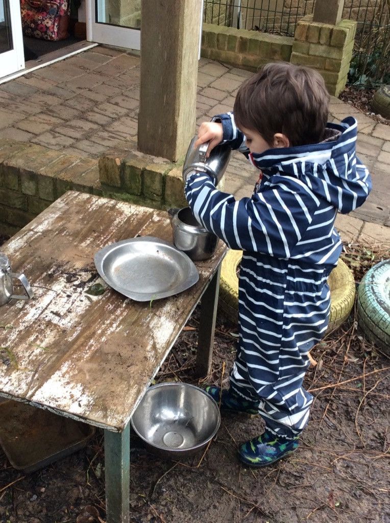 Young child pouring water into a metal pot from a steel thermos at an outdoor play area.