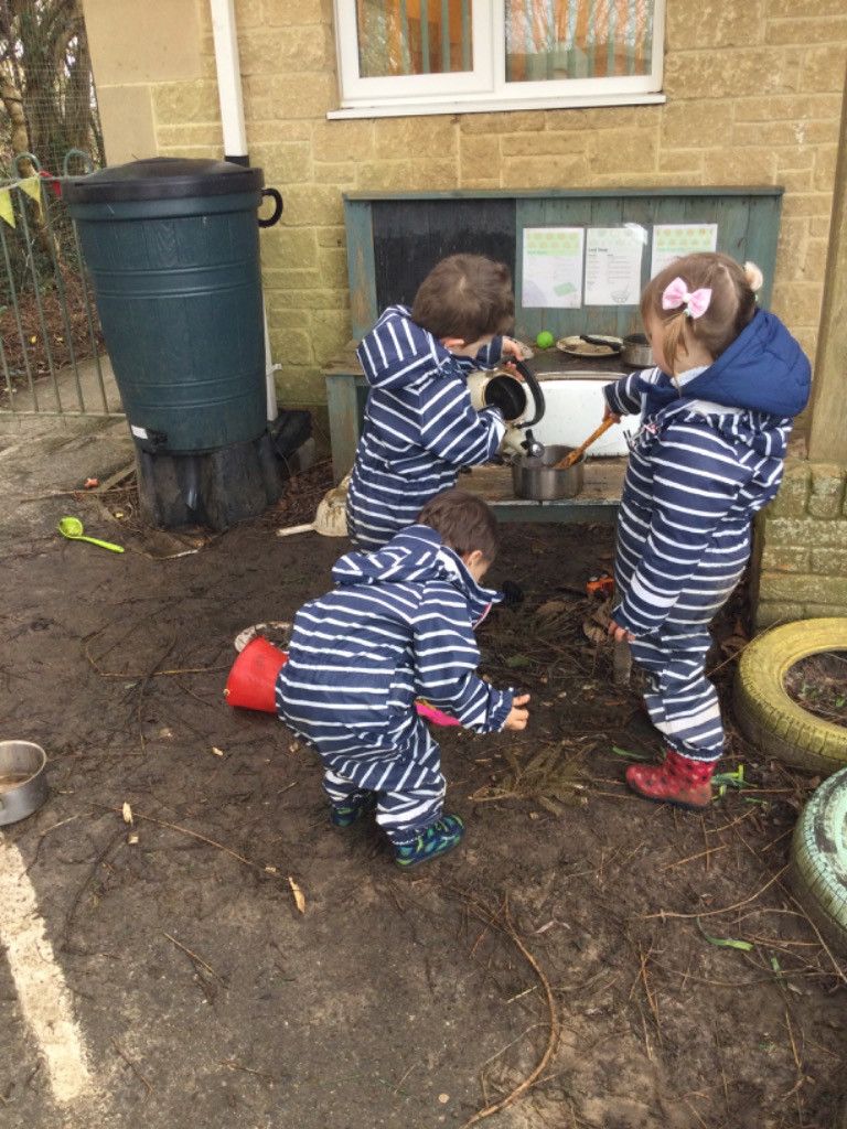 Three children in striped suits playing in mud near a water barrel, one pours from a kettle.