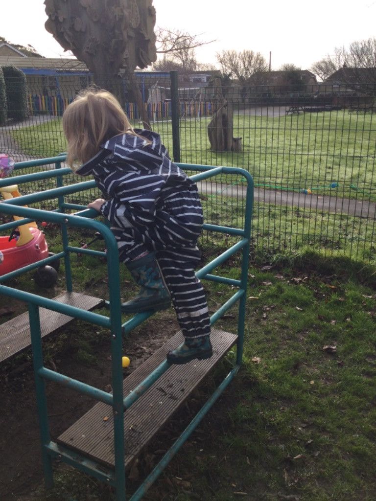 Child in striped rainsuit climbing a green metal play structure in a grassy outdoor setting.