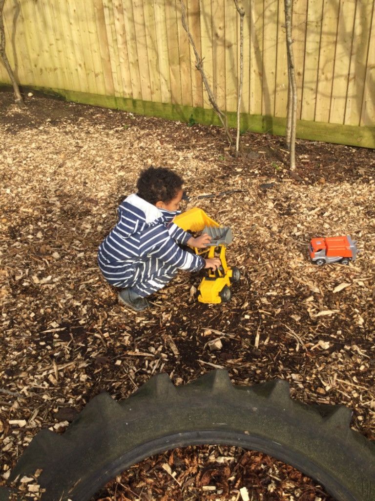 Young child plays with toy construction vehicles in a wood-chipped outdoor area.