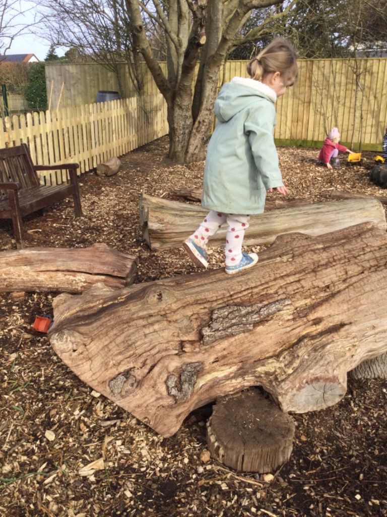 Child walking on a log in a play area. Another child plays nearby.