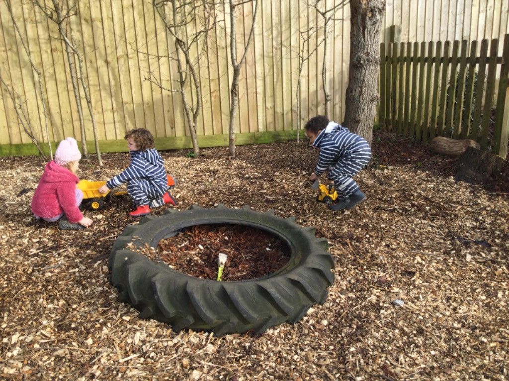 Three children playing with toy trucks in a wood-chipped outdoor area near a tire planter and a wooden fence.