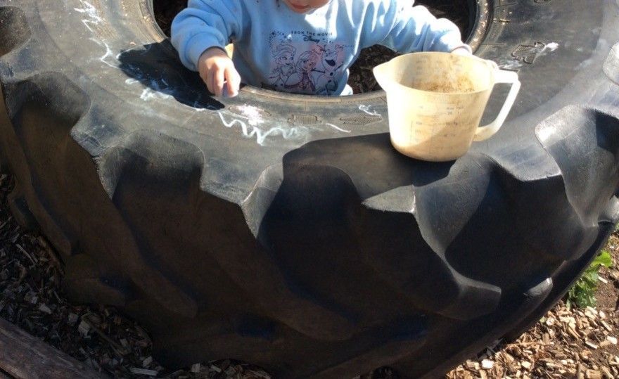 Child playing inside a large tire, drawing with chalk. A small cup sits nearby.