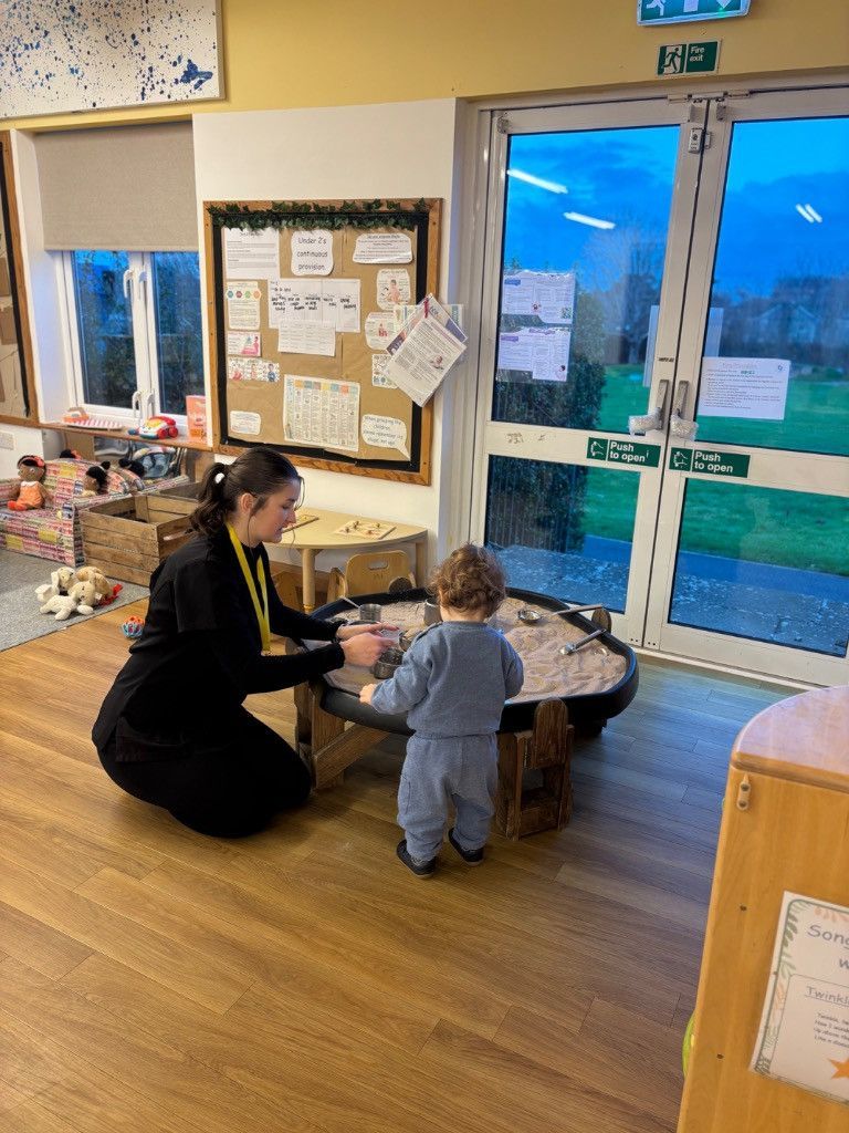Woman kneeling with toddler at sensory table; interior classroom with cloudy sky visible through doors.