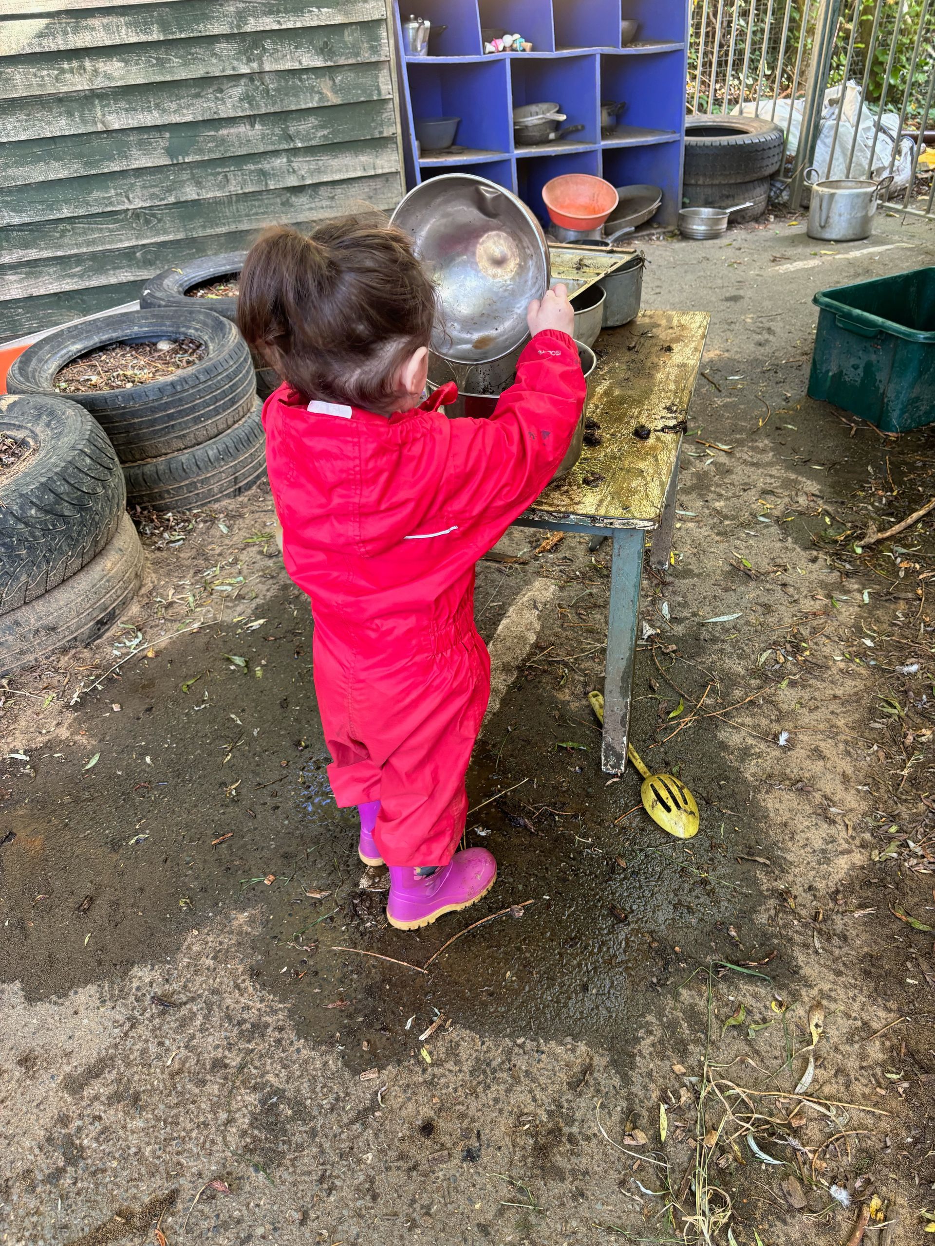 A little girl in a red jacket is playing in the mud with a bowl.