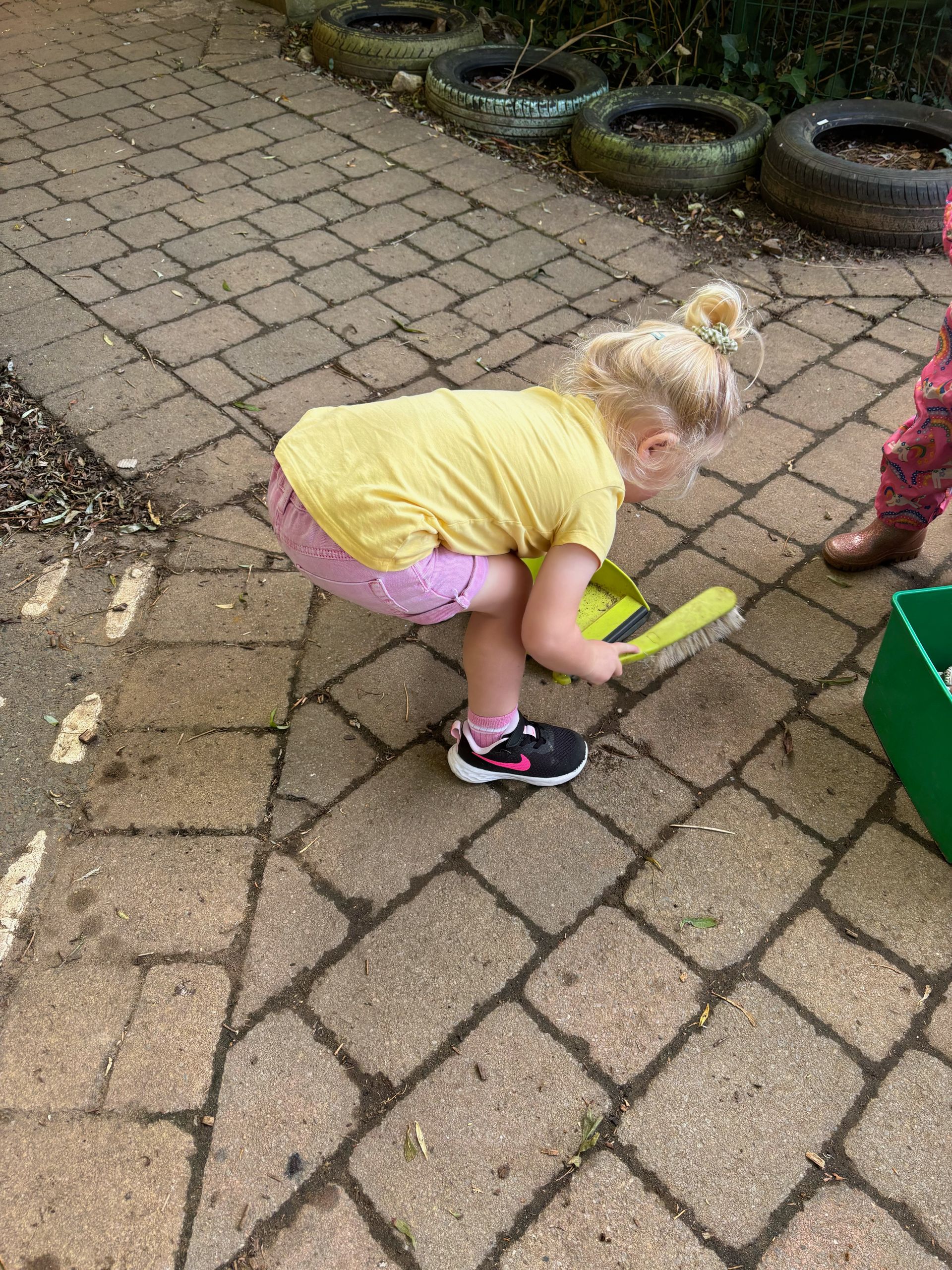 A little girl is playing with a broom on a brick sidewalk.