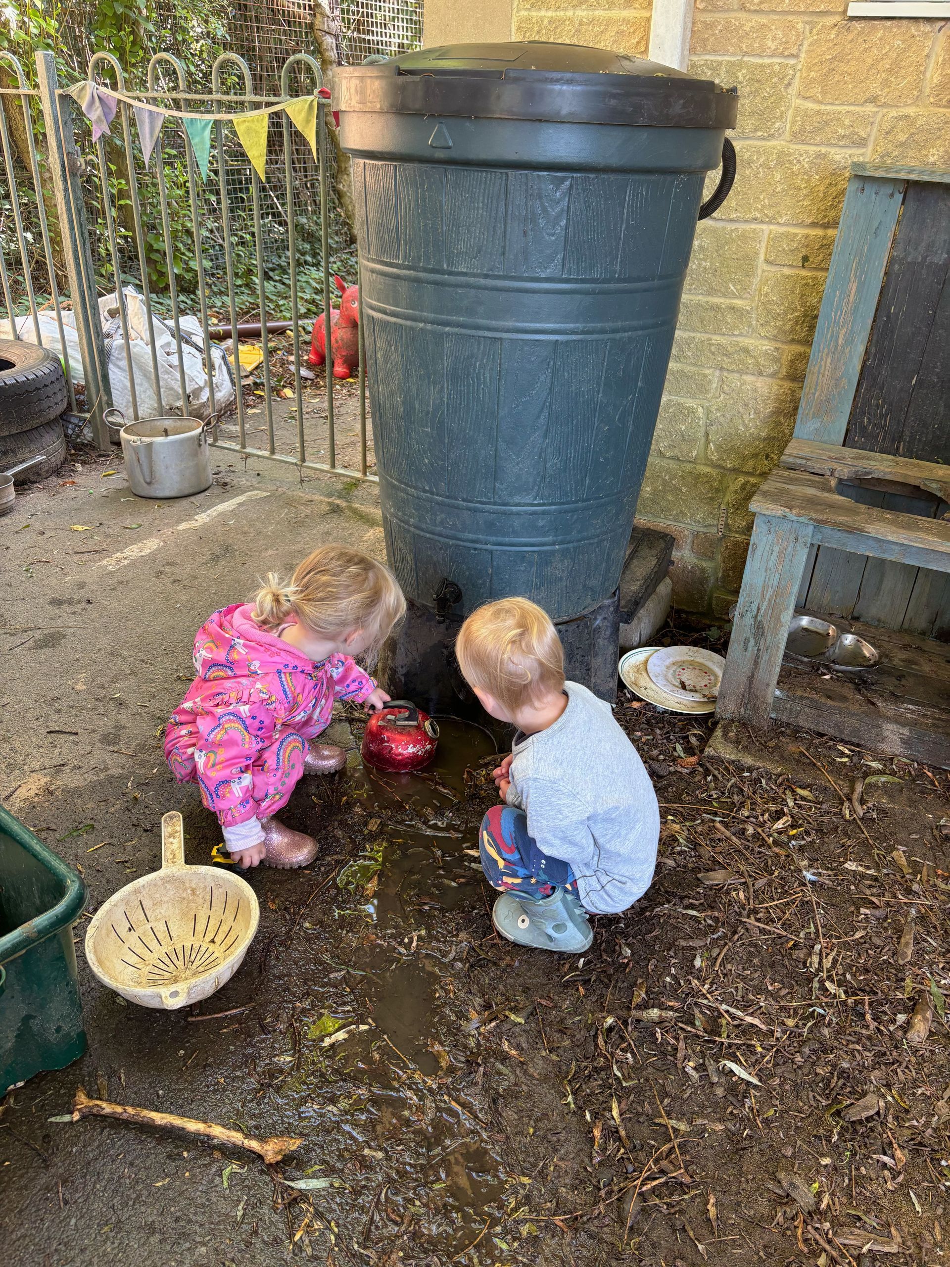 Two children are playing in the mud next to a large green barrel.