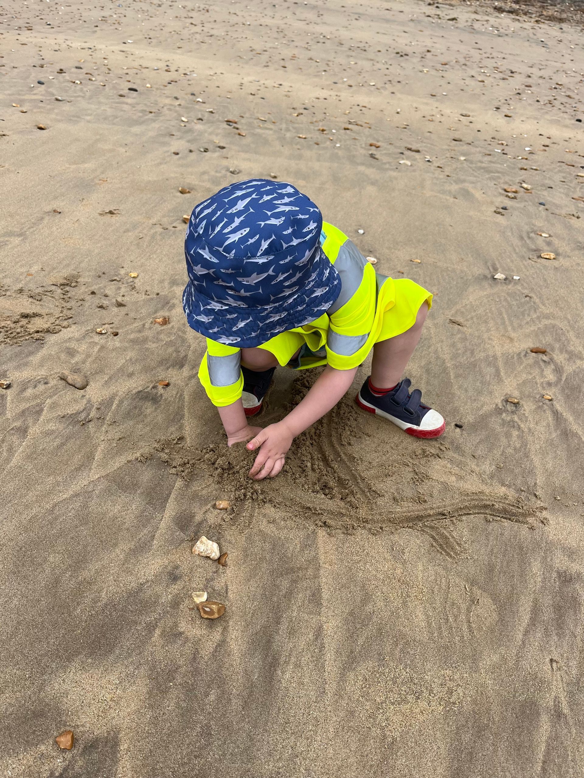 A little boy wearing a hat and safety vest is playing in the sand.