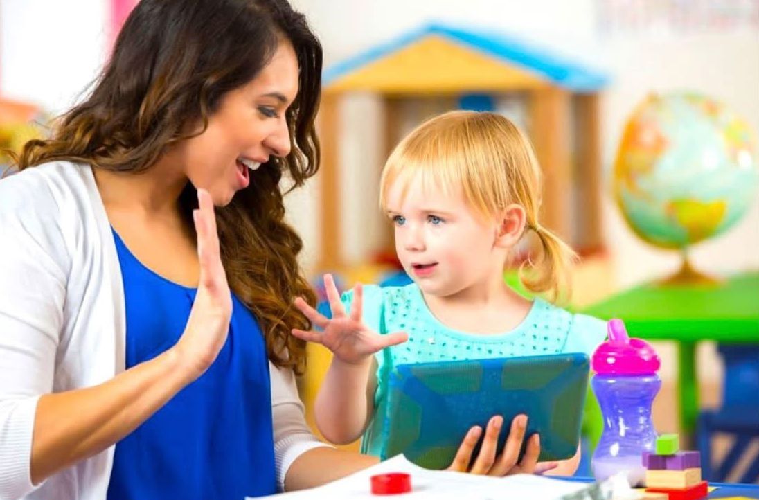 Woman and young child painting at a classroom table, smiling and showing colorful handprints