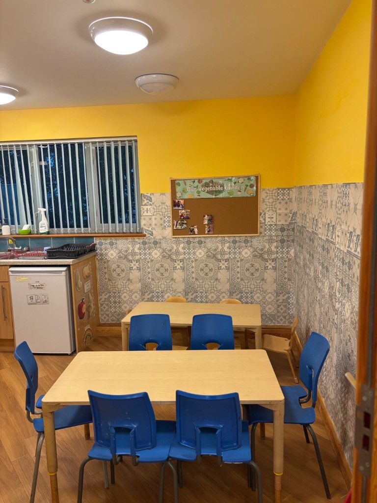 A playroom with yellow and patterned walls, tables, blue chairs, and a fridge.