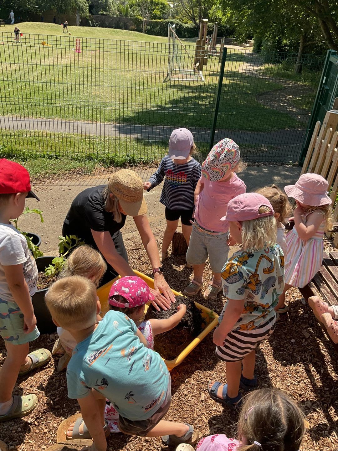Children gather around a small garden bed outdoors, planting or digging in soil with tools.