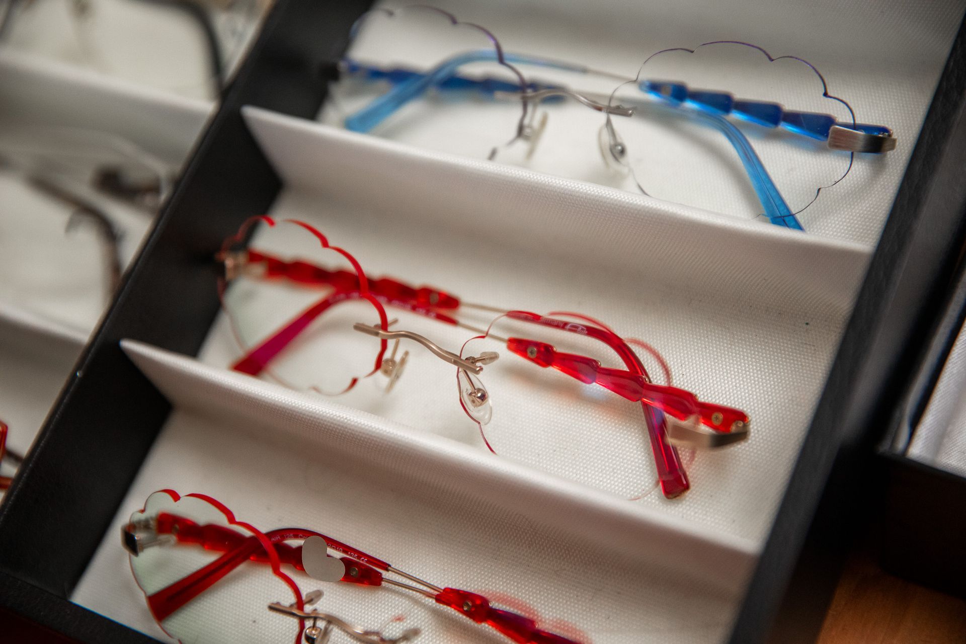 White display tray holding several red and blue pairs of eyeglasses/cases on fabric-lined shelves