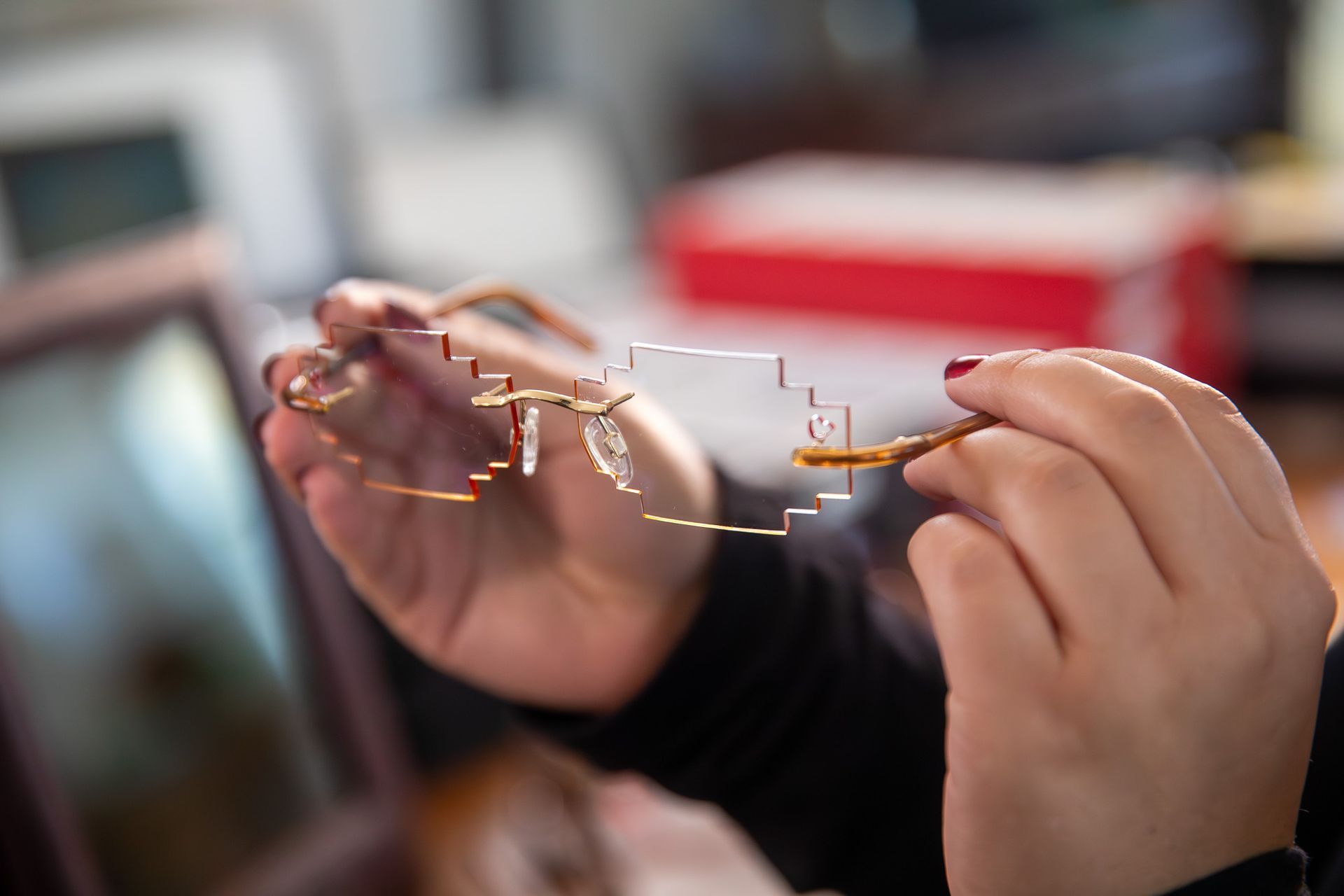 Hands holding a pair of eyeglasses with a small wire repair tool, close-up on a blurred indoor background
