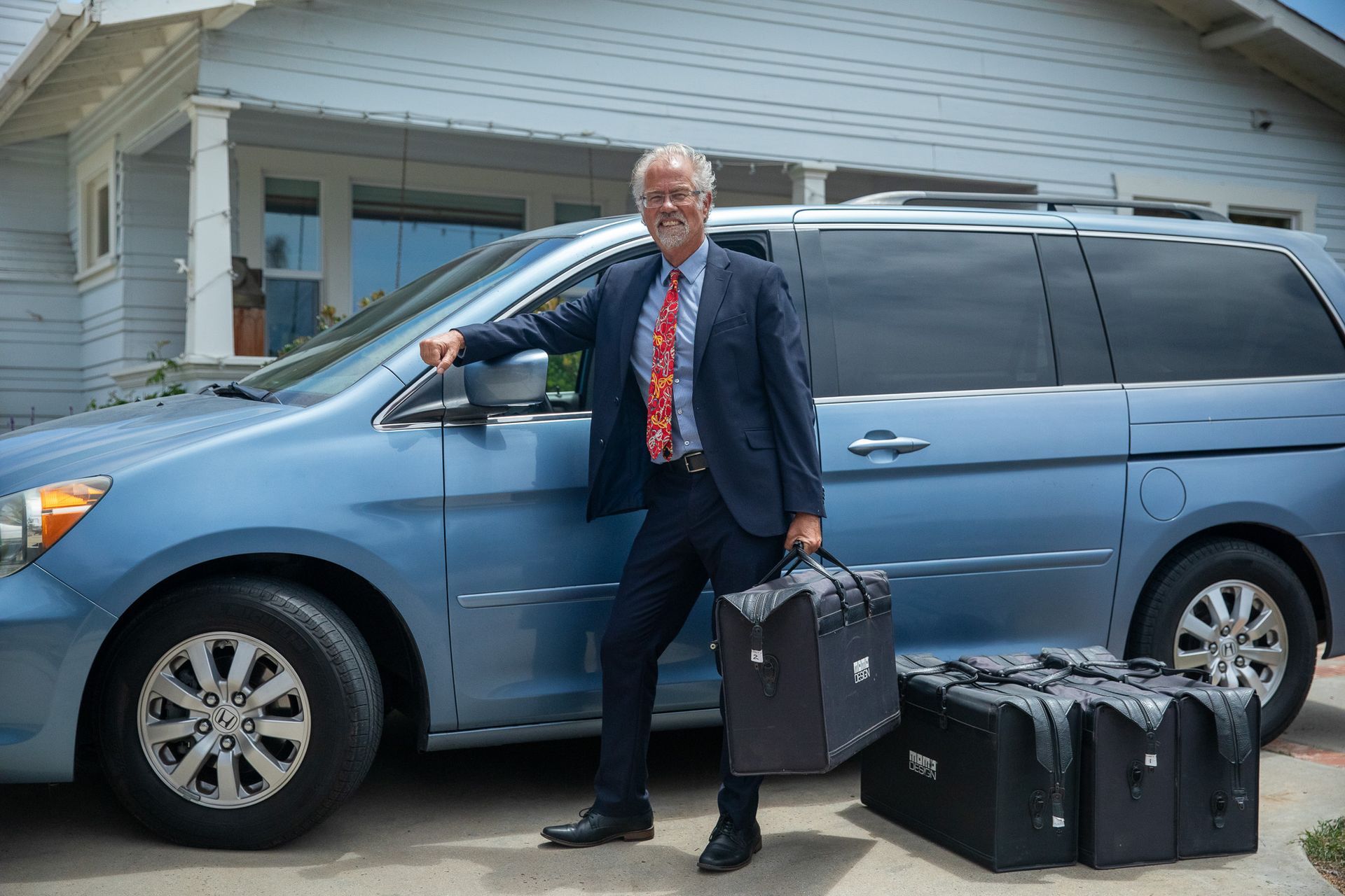Man standing by a blue minivan with luggage on the driveway in front of a house