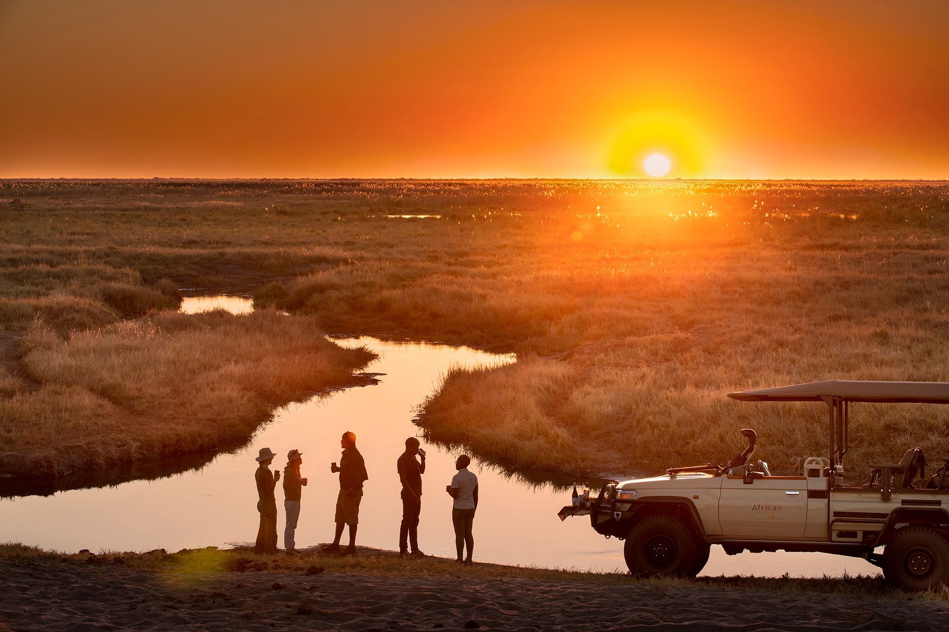 Silhouetted people stand by a winding river during a sunset in a grassy savanna with a safari vehicle parked nearby.
