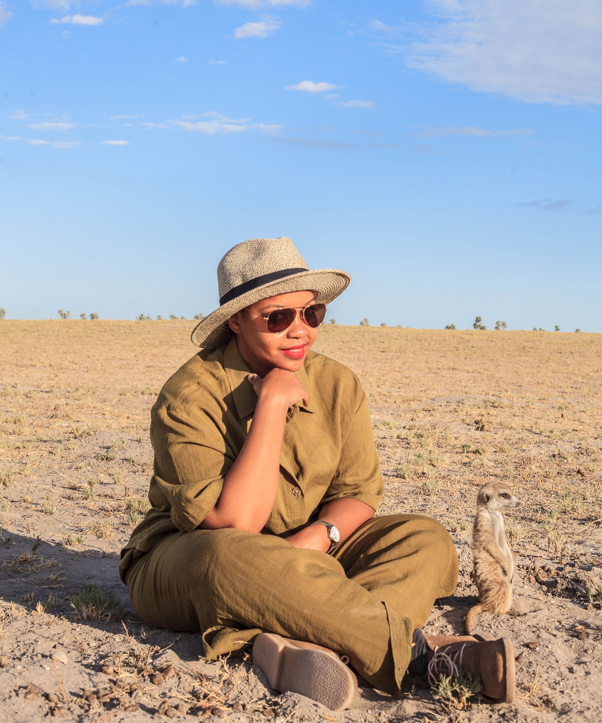A person in a tan hat and outfit sits cross-legged on dry, sparse ground next to a small meerkat under a bright blue sky.