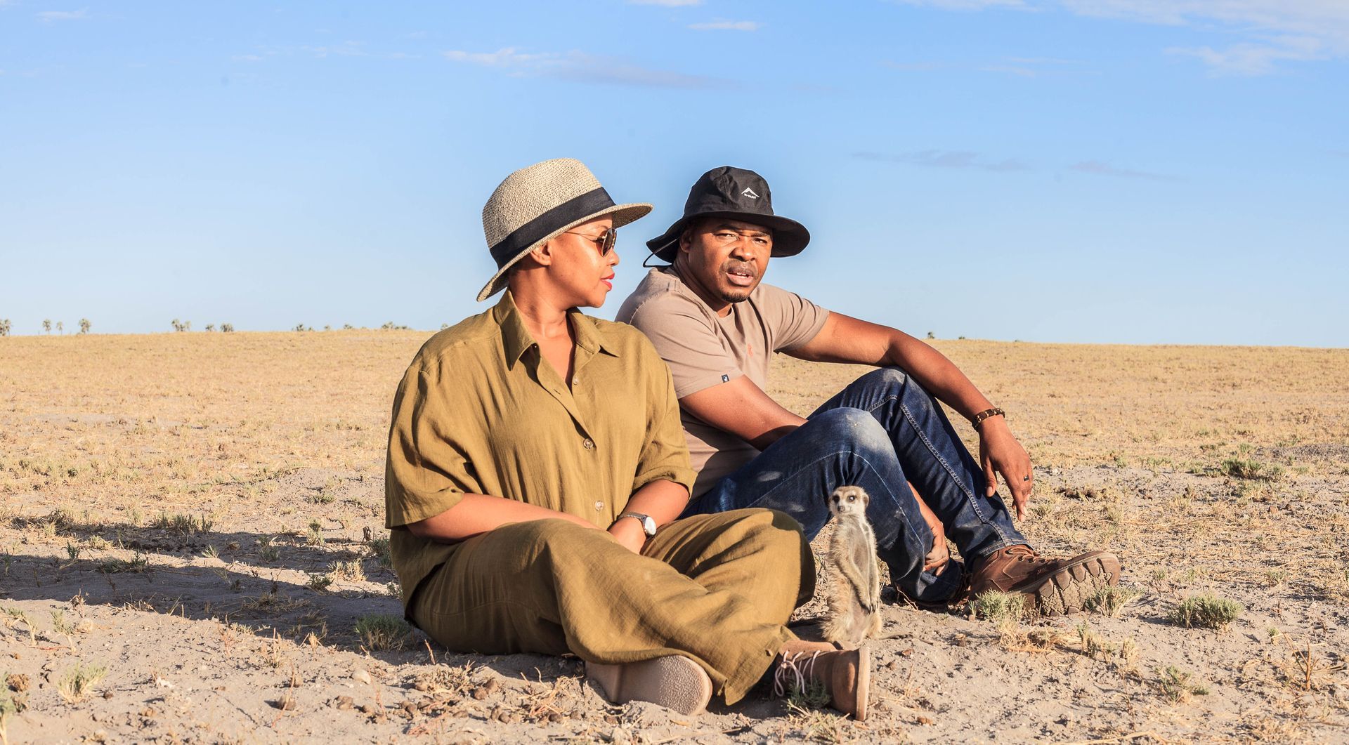 Two people in safari hats sit on the ground in a sparse, arid landscape, observing a small animal nearby.
