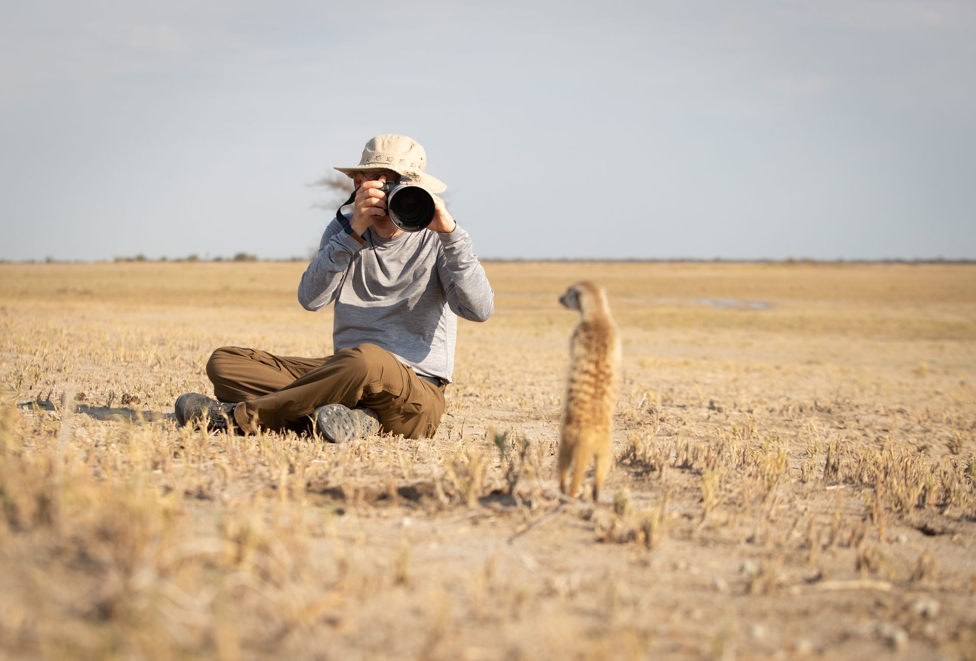 A photographer sits in a dry, open field, aiming a long camera lens at a meerkat standing upright nearby.