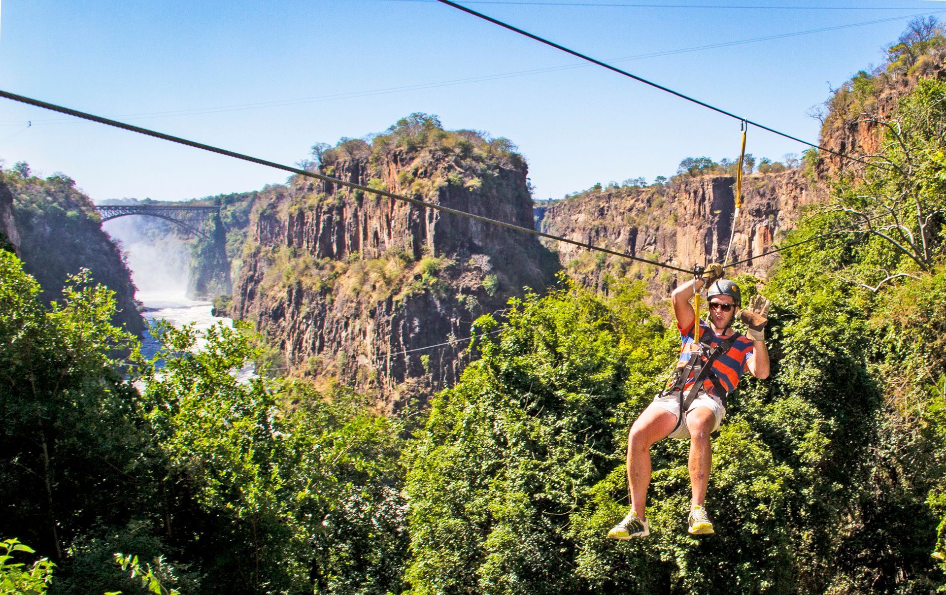 A person wearing a safety harness ziplines across a deep gorge near a misty waterfall and a high bridge.