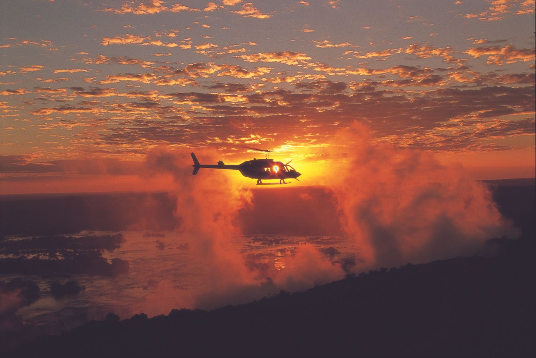A helicopter flies through a misty landscape at sunset, silhouetted against a bright, orange-glowing horizon.