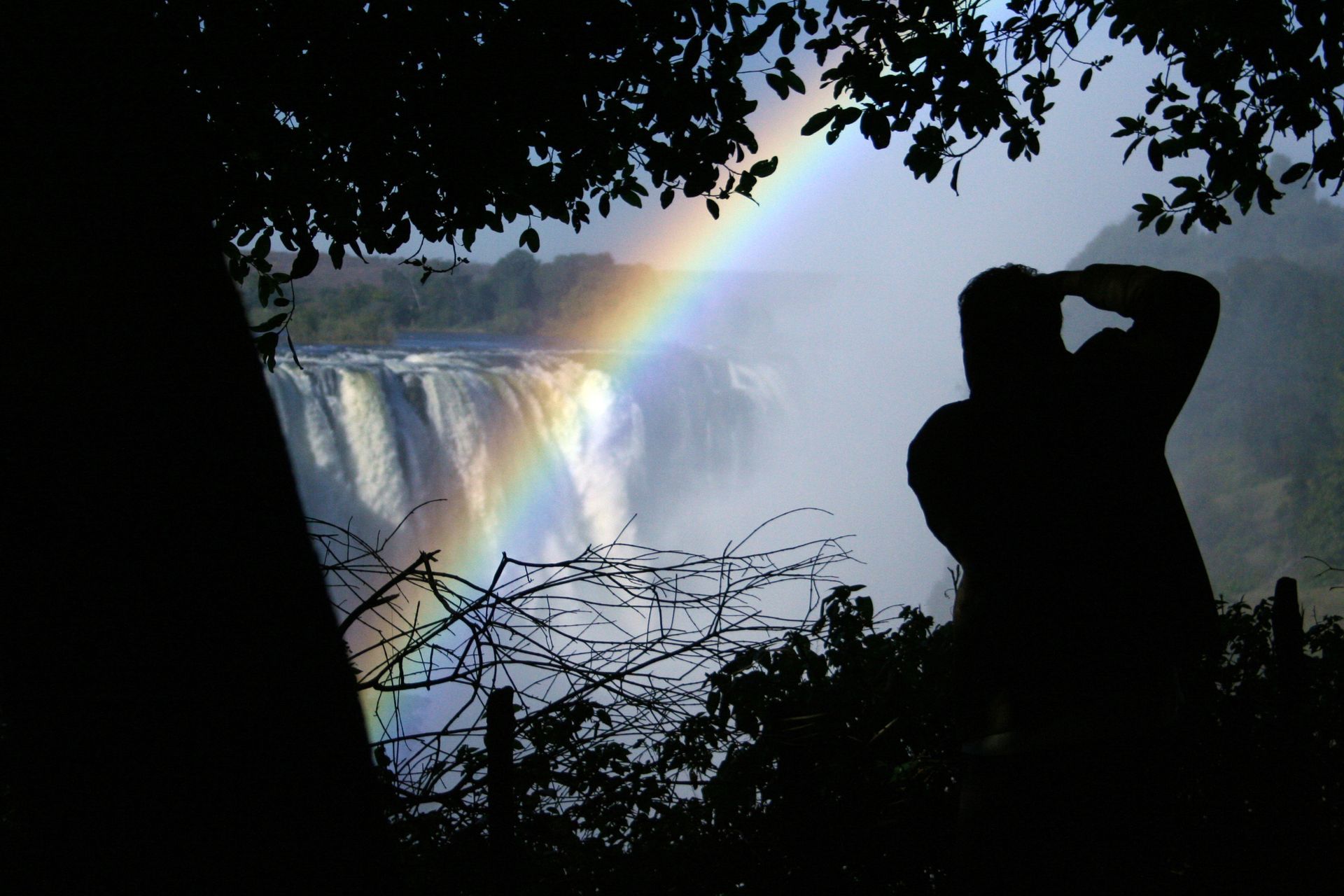 A silhouette of a person viewing a rainbow over a large waterfall, framed by tree branches in the foreground.