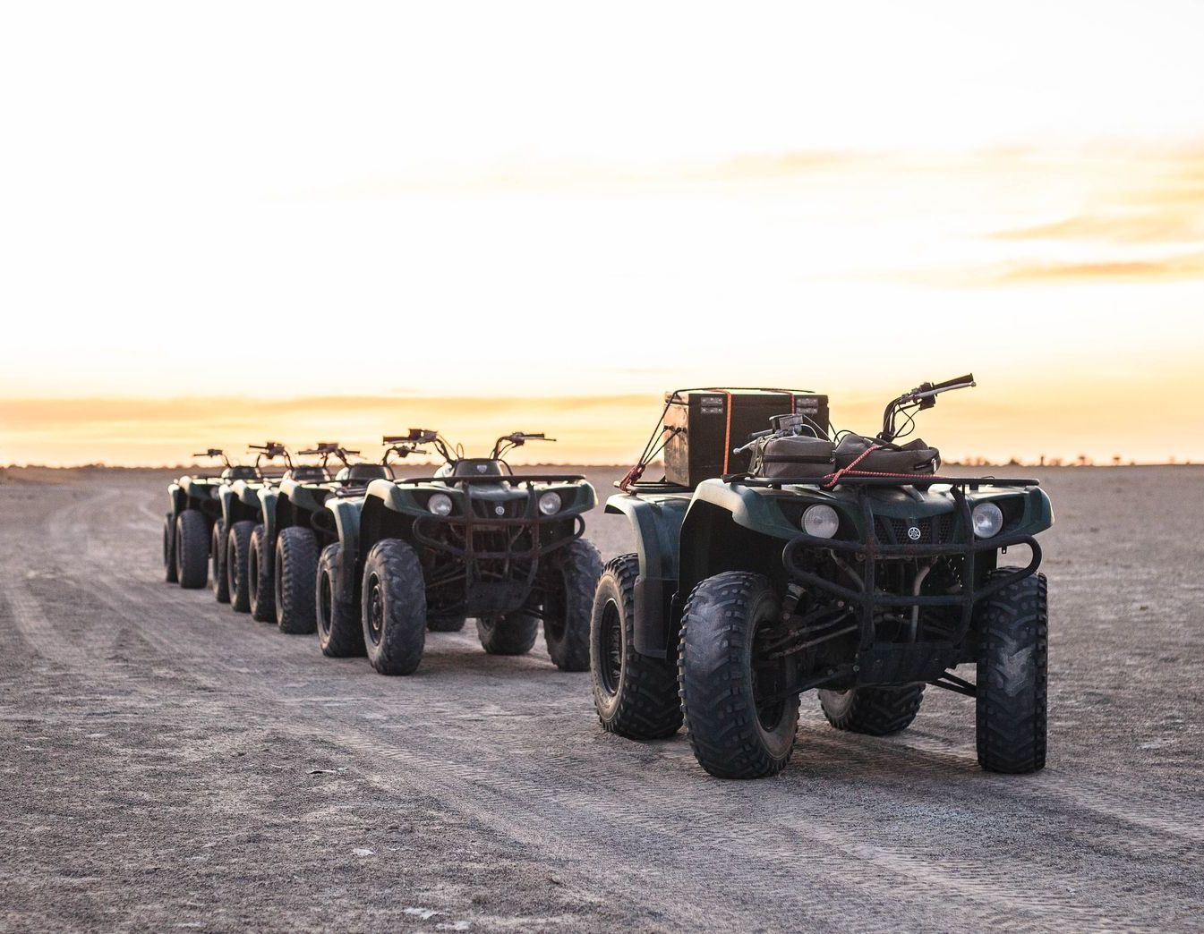 A line of dark green ATVs parked on a dirt trail under a sunset sky.