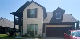 A two-story house with cream-colored brick, dark wood trim, a front balcony, a dormer window, and a red car parked outside.