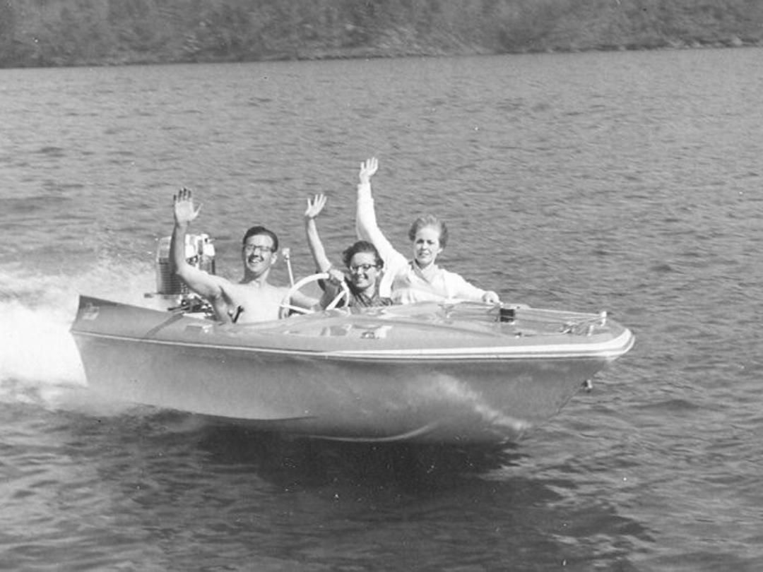 People in a bass boat wave on a lake, water splashing.