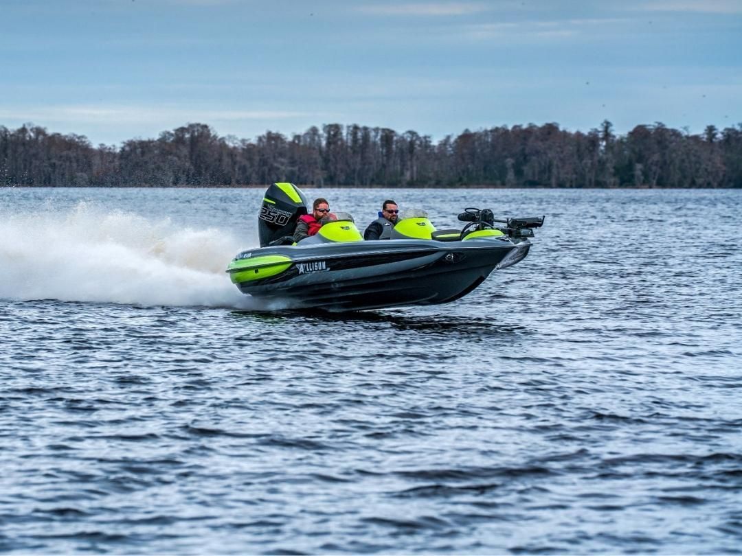 Speedboat on a lake, splashing water. Two people in the boat, overcast sky, trees in the background.