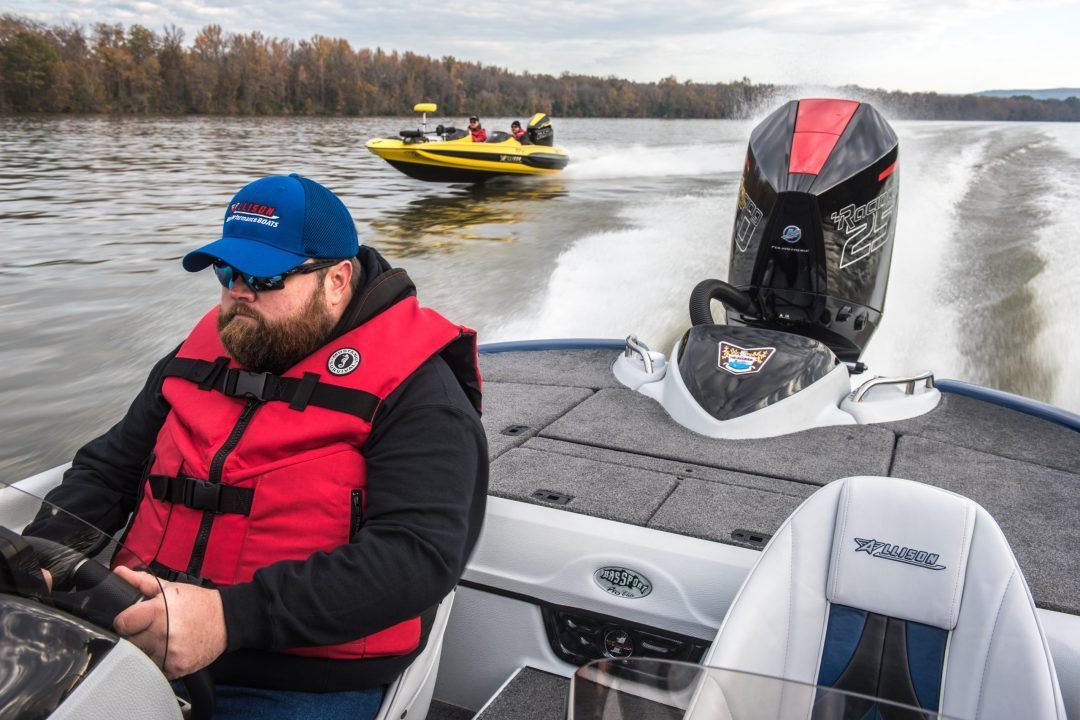 Man piloting a bass boat on a lake, wearing a life vest. A yellow boat follows in the distance.