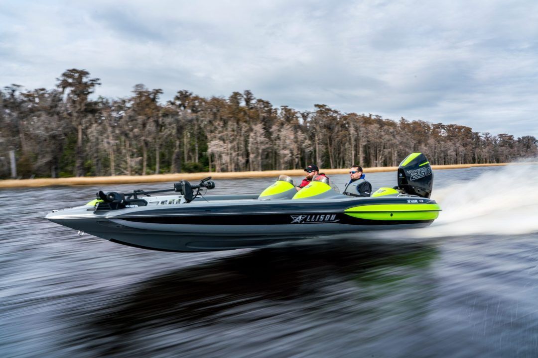 Fishing bass boat speeding across water, two people on board, gray boat with neon green accents, trees in the background.