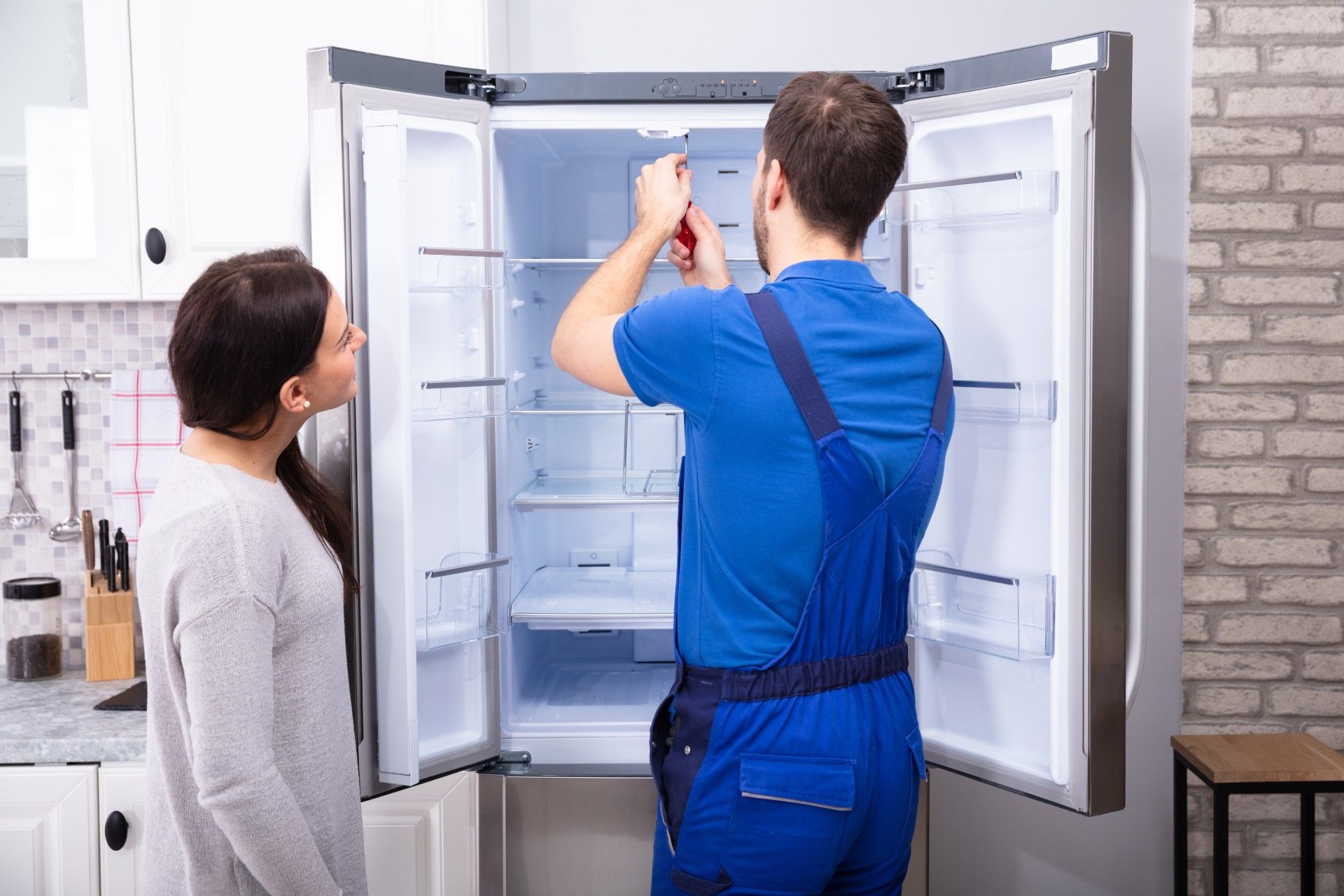 A man is fixing a refrigerator in a kitchen while a woman looks on.