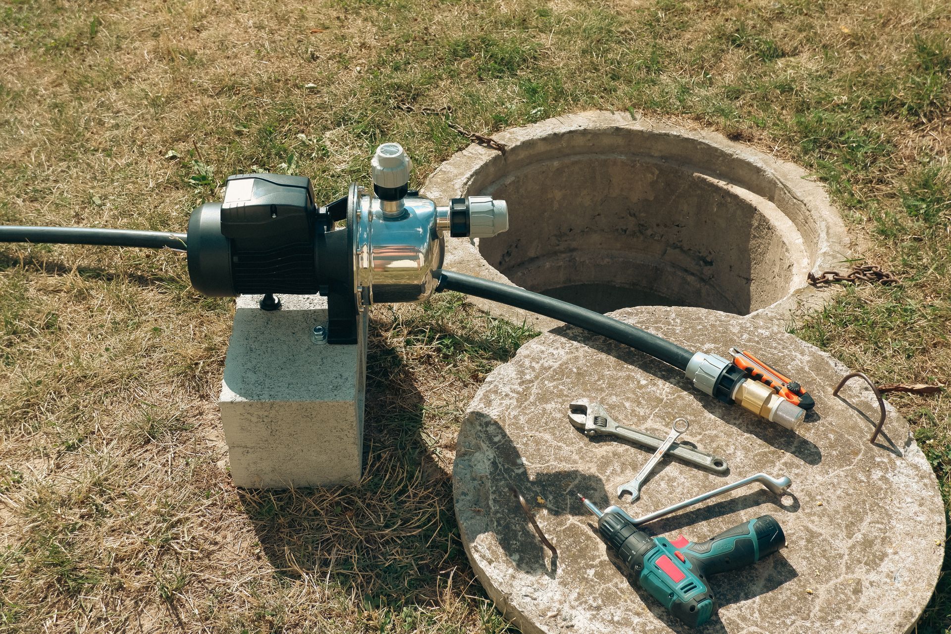 Outdoor water pump beside an open utility pit, with tools on a round concrete cover nearby.