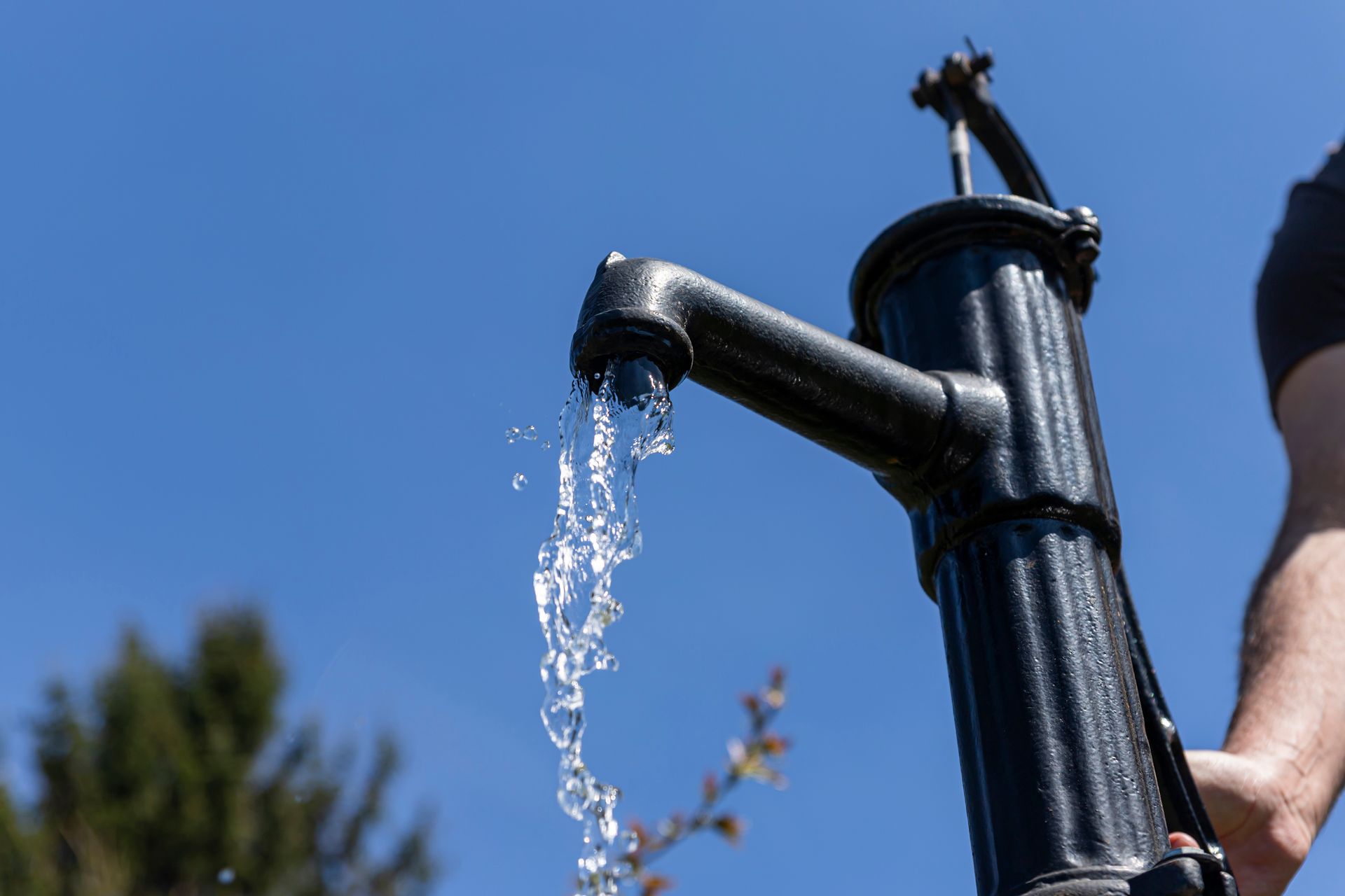 Close-up of a black hand-operated well pump spout releasing clear water against a bright blue sky.