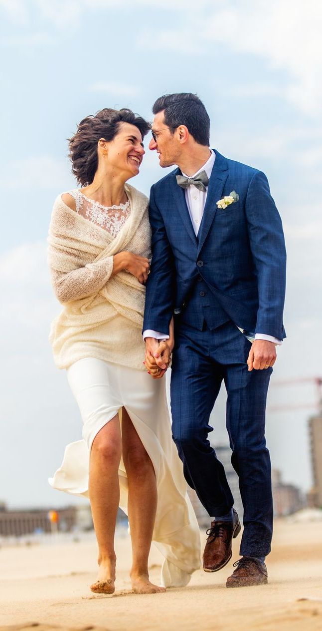 Een stel in bruidsjurken loopt hand in hand, lachend, over het strand. De bruidegom draagt ​​een blauw pak, de bruid een witte jurk en een beige sjaal.