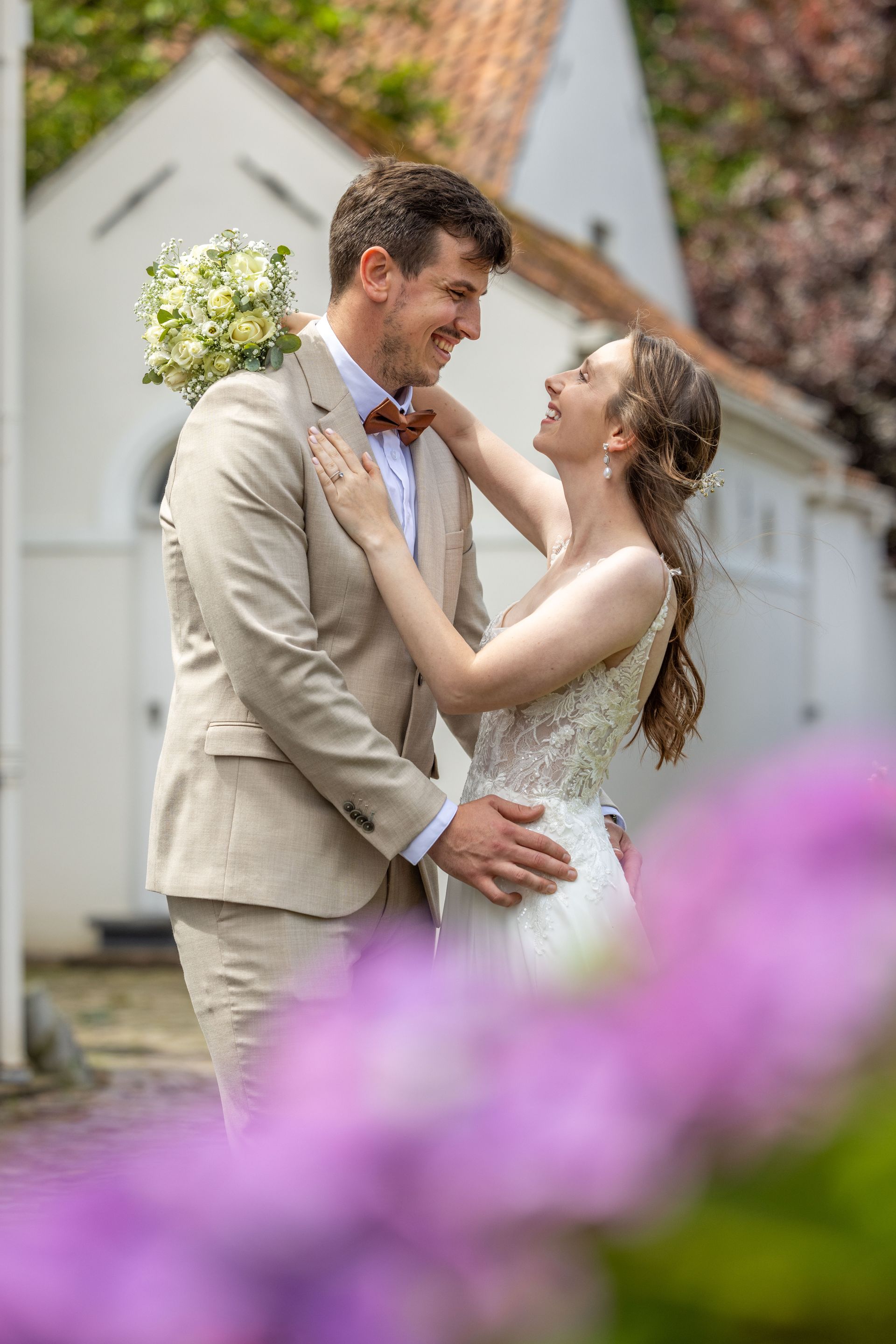 Bruid en bruidegom omhelzen elkaar en kijken elkaar aan, voor een wit gebouw, omringd door paarse bloemen.