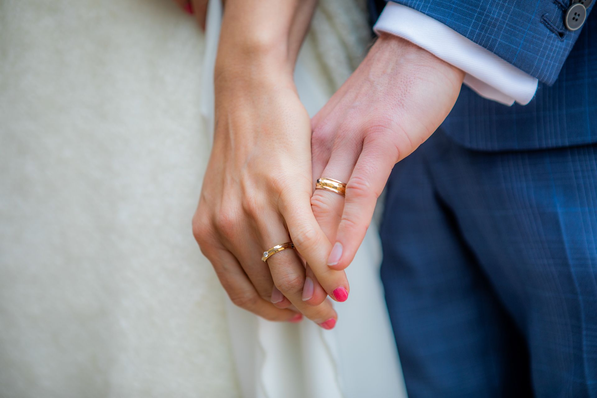 Een stel houdt elkaars hand vast en draagt ​​trouwringen; de vrouw draagt ​​een witte jurk, de man een blauw pak.