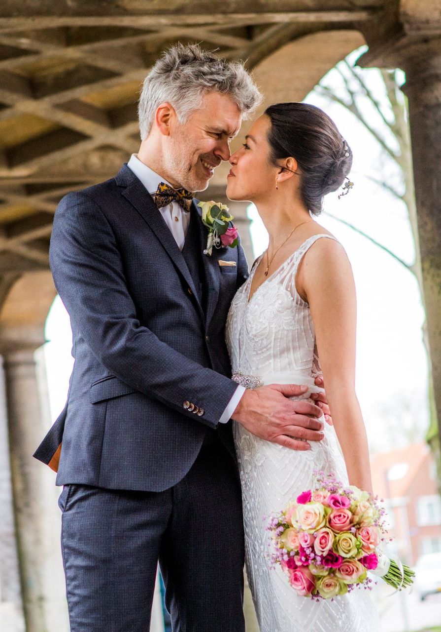 Bruidspaar omhelst elkaar, dicht bij elkaar, onder een boogconstructie, glimlachend. Bruid in witte jurk, bruidegom in pak.