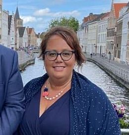 Een vrouw met een bril en een blauwe sjaal lacht voor een kanaal in Brugge, België.