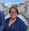Een vrouw met een bril en een blauwe sjaal lacht voor een kanaal in Brugge, België.