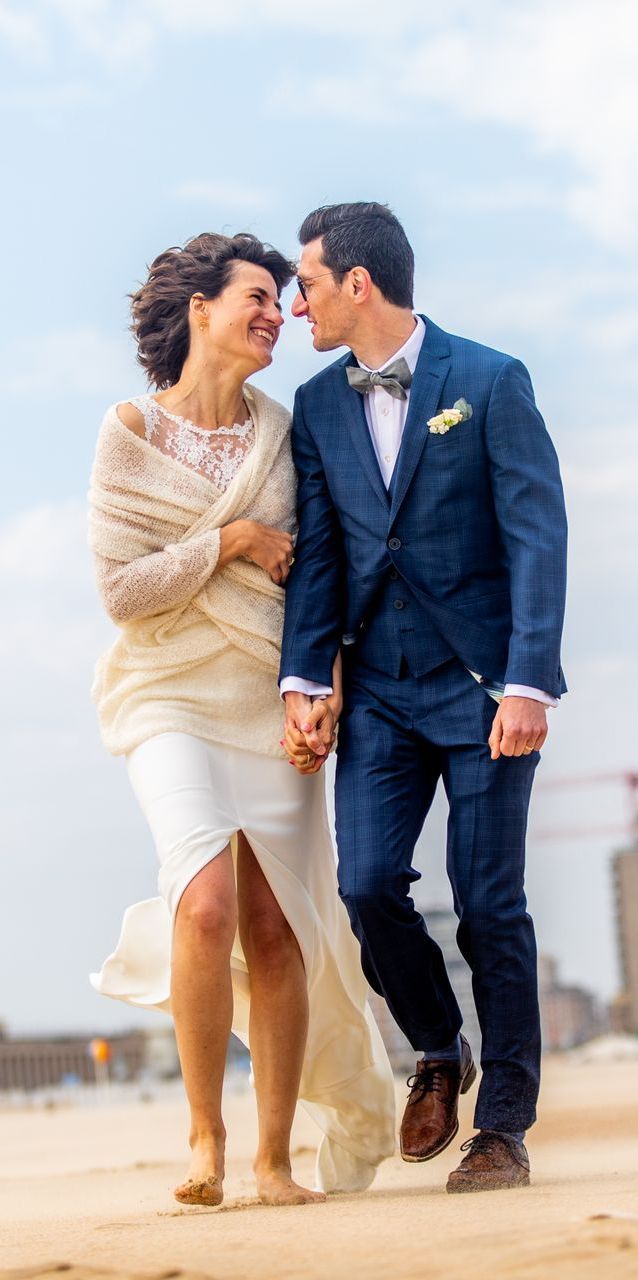 Een stel loopt hand in hand op het strand; glimlachend kijkend naar elkaar. De man draagt ​​een blauw pak, de vrouw een trouwjurk.