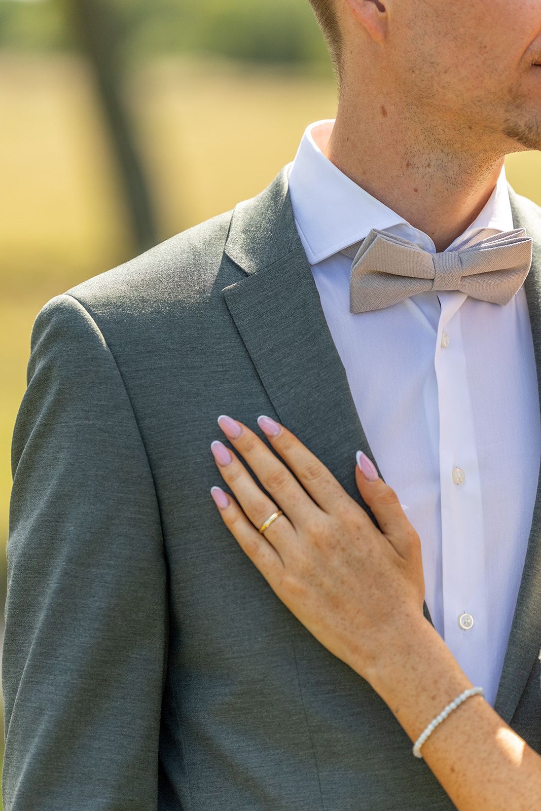 De hand van een vrouw op de borst van een man; beiden formeel gekleed. Zachtroze nagels, grijze blazer en vlinderdas. Buiten, zonnig.