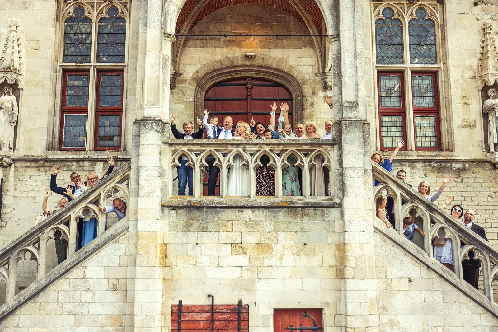 Mensen zwaaien op een balkon, een trap voor een stenen gebouw met boogramen, een bruiloftsfeest.