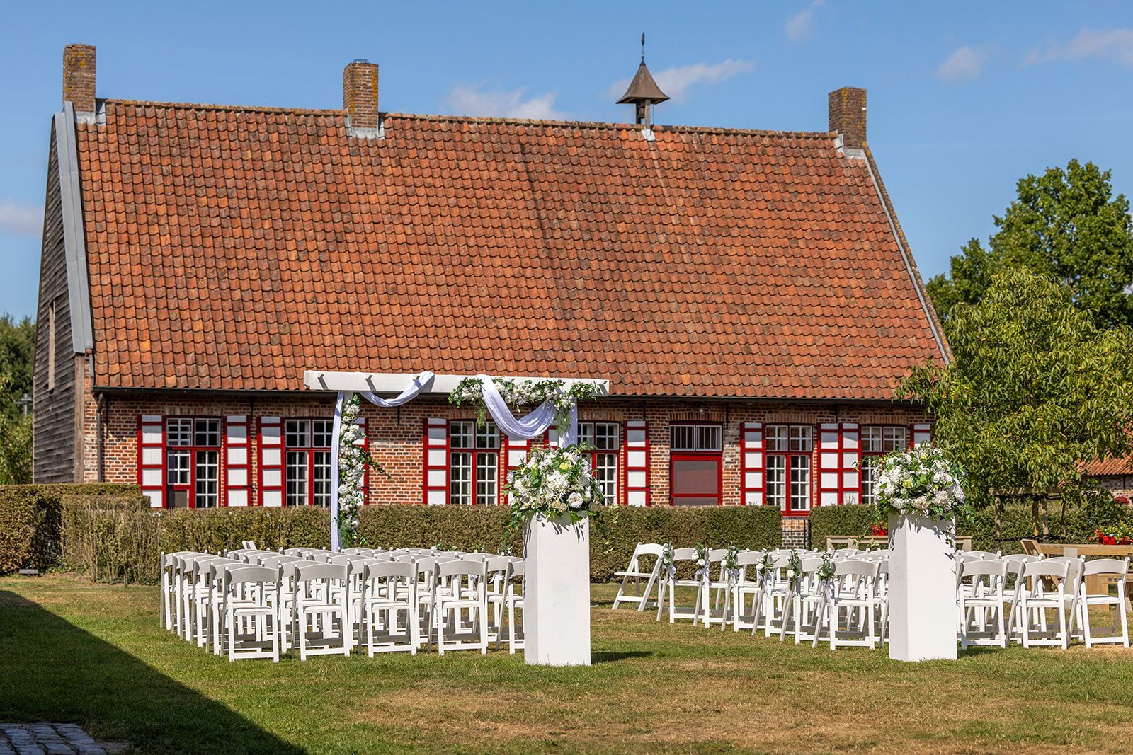 Huwelijksceremonie met witte stoelen, bloemen en een versierde boog voor een gebouw van rode baksteen.