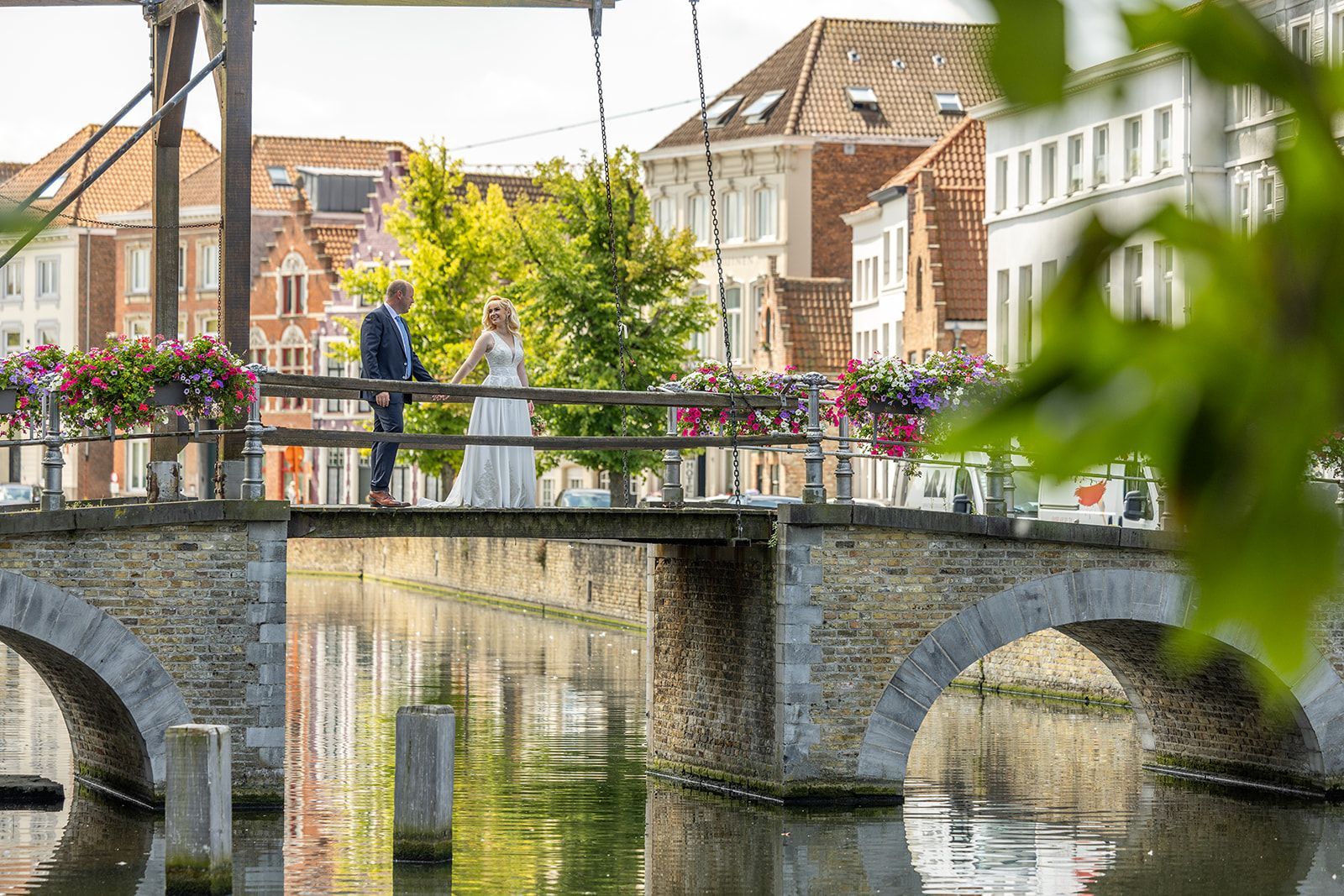 Paar op stenen brug over kanaal, gebouwen op de achtergrond, bloemenmanden.