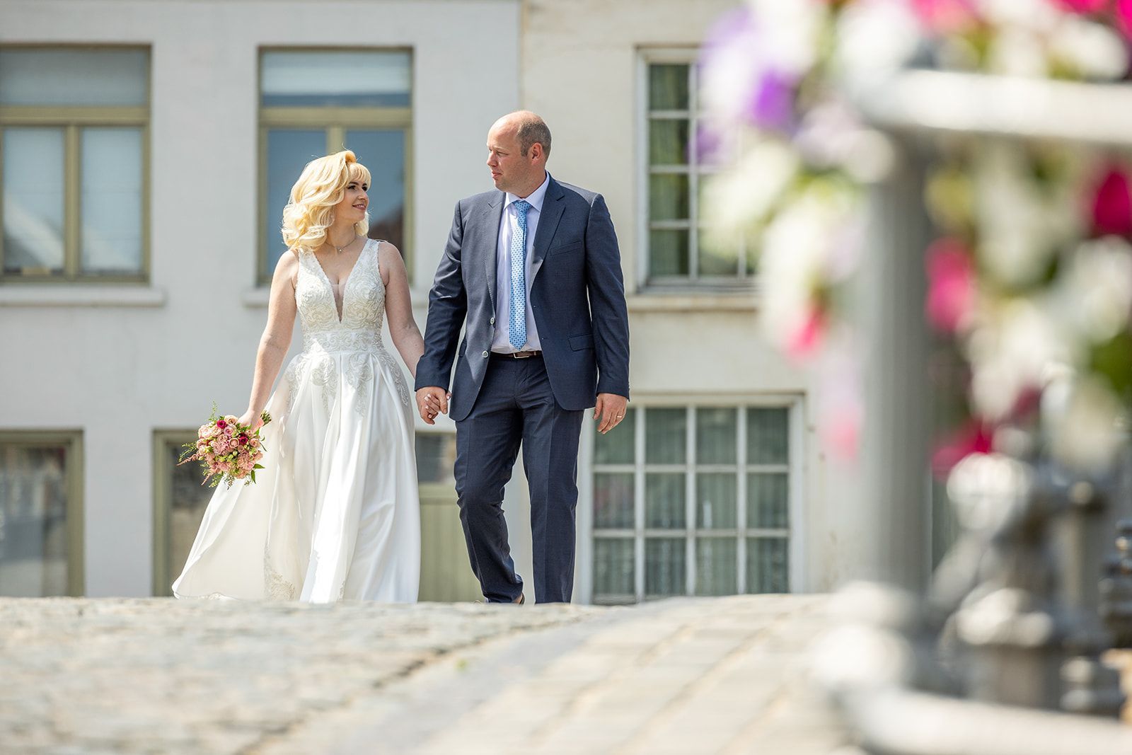 Bruid en bruidegom lopen hand in hand buiten, in witte jurk en pak. Gebouwen op de achtergrond, bloemen op de voorgrond.
