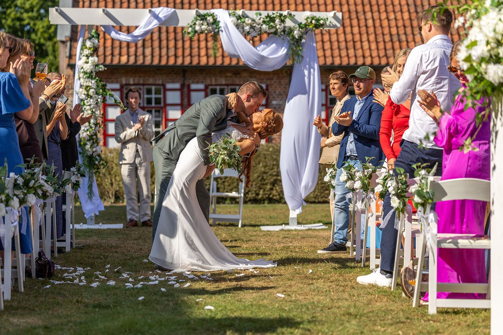 Een kussend stel na de huwelijksceremonie; klappende gasten. Een witte boog, bloemendecoraties en een zonnige buitenomgeving.