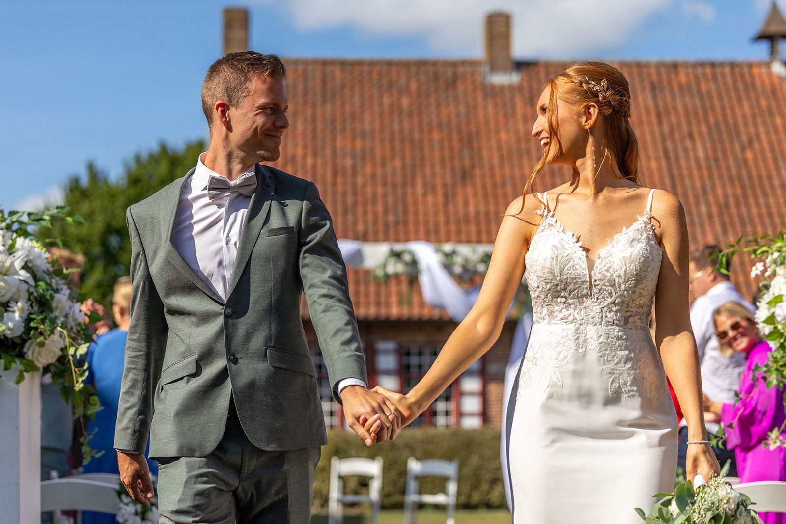 Een stel in bruidsjurk loopt hand in hand en lacht naar elkaar, buiten. Op de achtergrond een gebouw met een rood dak.
