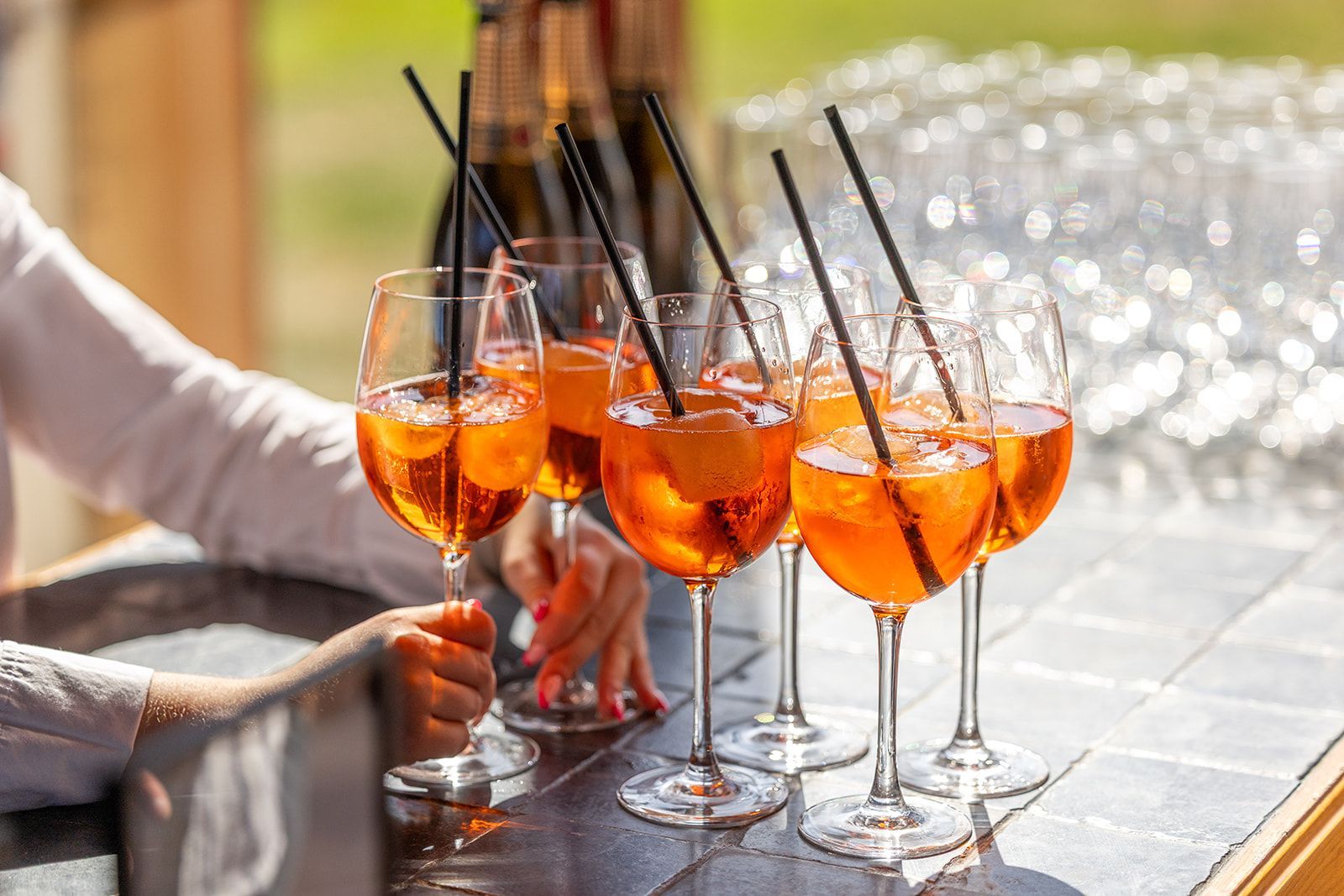 Persoon die buiten een dienblad met Aperol Spritz-cocktails vasthoudt, met zwarte rietjes.